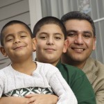 Latino father and two young sons sitting together in front of a house