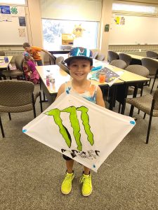 Cloverbud wearing a tie-dyed shirt, a blue hat, and yellow shoes showing their Monster kite they made.
